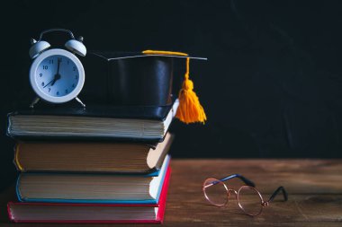 Masaüstünde kitaplar, yazma malzemeleri, gözlükler, defter ve çalar saat var. Siyah arka plan. Öğrenme ve kendini eğitme konsepti. Seçici odak.