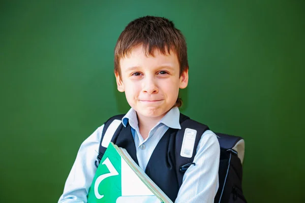 Funny schoolboy grimaces near the green school board in the classroom. Elementary school child with bag and books. Back to school.