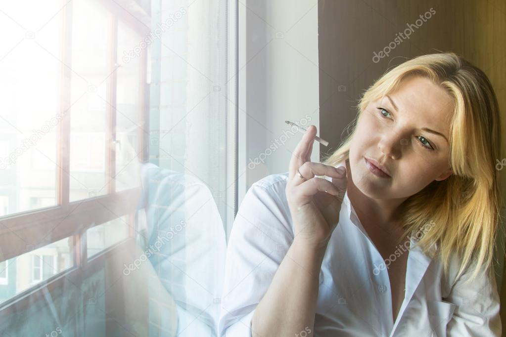 The woman smokes looking out the window. 