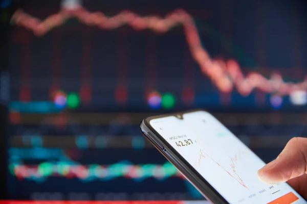 Person checking the stock prices on a smartphone in front of a big screen market rates. Person viewing share prices on a smartphone with market prices on a screen in the background.