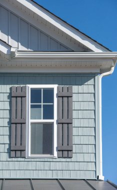 Close up view of classic double hung sash window with wooden shutters on a newly build house with green shake and shingle siding , white gutter system in America blue sky clipart