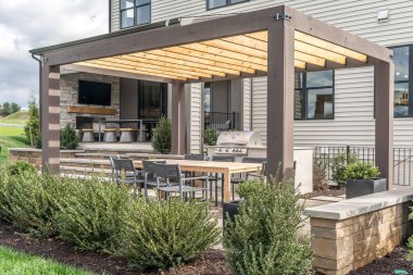 Açık hava pergola gölgelik yapısı, tente ve teras çatısı, yemek masası, sandalyeler, bahçe düzenleme ile çevrili metal ızgara.
