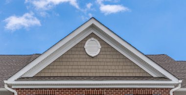 Aerial view of a double gable roof with triangle gable decoration and elegant colonial white round gable vent on a beige shake and shingle siding roof on a luxury town home with blue cloudy sky clipart