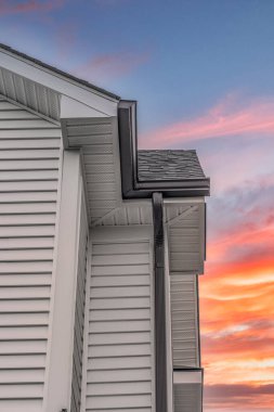 Closeup view of dark metal gutter system on white horizontal vinyl siding, fascia, soffit, on a pitched roof attic at a luxury American single family home dramatic colorful sunset sky background clipart