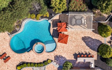 Turkuaz su, beyaz açık teras pergola, oturma, yemek masası, turuncu plaj sandalyeleri, eğimli peyzaj, turuncu plaj şemsiyeleri olan havadan manzaralı havuz.