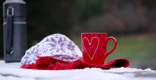 a red mug with a heart stands on a table with a red towel and a thermos on a blurred background