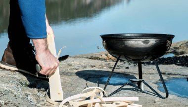 Bir adam doğada, nehrin kıyısında, taşların üzerinde barbekü yakıyor.