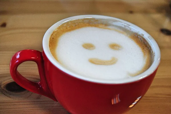 Czerwona filiżanka kawy latte z uśmiechniętym rysunkiem twarzy w pianie dla leworęcznych. — Zdjęcie stockowe