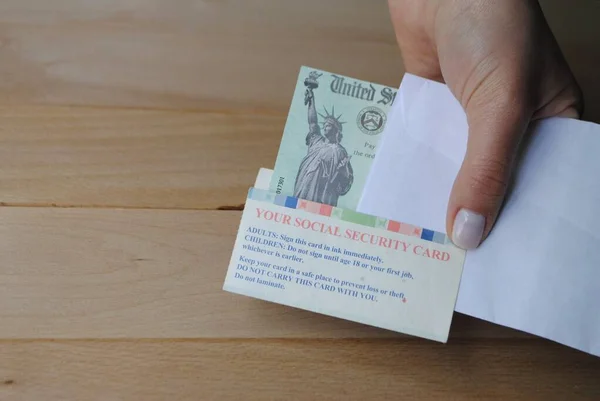 Hand holds the US federal stimulus check showing the statue of liberty with envelop next to social security card. SSN holders receive the Covid 19 pandemic relief Economic Impact Payments.