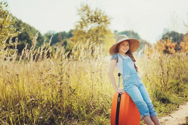 Glückliches Mädchen mit orangefarbenem Koffer auf Sommerurlaub. Kinder gehen aufs Land. Gemütliche ländliche Szenerie — Stockfoto