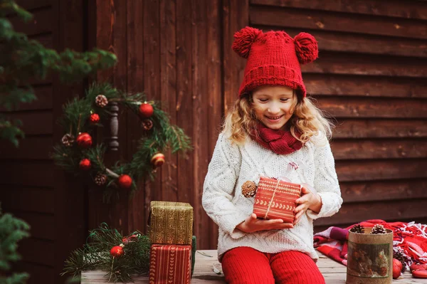 Happy child girl in red hat and scarf wrapping Christmas gifts at cozy country house, decorated for New Year and Christmas — Stock Photo, Image