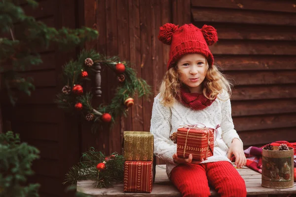 Happy child girl in red hat and scarf wrapping Christmas gifts at cozy country house, decorated for New Year and Christmas — Stock Photo, Image