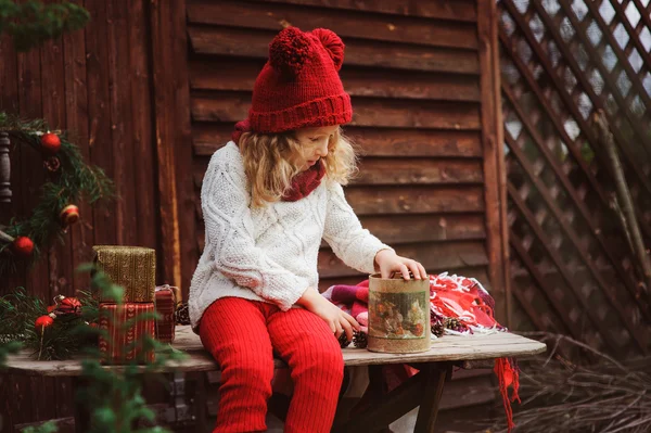 Happy child girl in red hat and scarf wrapping Christmas gifts at cozy country house, decorated for New Year and Christmas — Stock Photo, Image