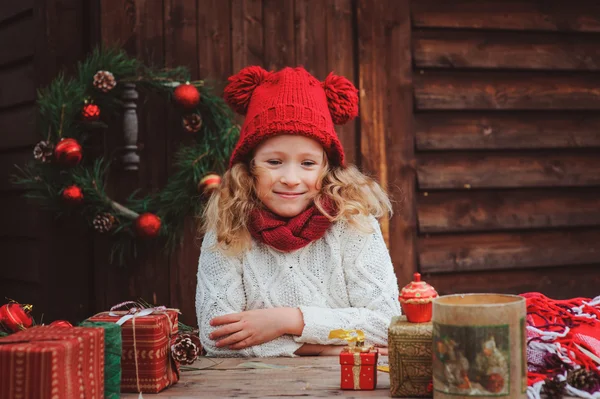 Happy child girl in red hat and scarf wrapping Christmas gifts at cozy country house, decorated for New Year and Christmas — Stock Photo, Image