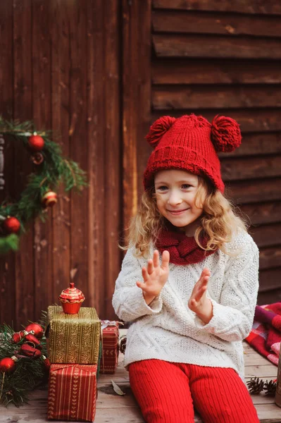 Happy child girl in red hat and scarf wrapping Christmas gifts at cozy country house, decorated for New Year and Christmas — Stock Photo, Image