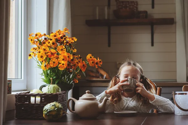 Niña desayunando en casa en otoño por la mañana. La vida real acogedor interior moderno en casa de campo. Niño comiendo rosquillas y bebiendo té . — Foto de Stock