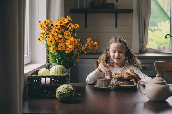Niña desayunando en casa en otoño por la mañana. La vida real acogedor interior moderno en casa de campo. Niño comiendo rosquillas y bebiendo té . — Foto de Stock