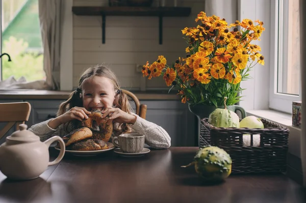 Niña desayunando en casa en otoño por la mañana. La vida real acogedor interior moderno en casa de campo. Niño comiendo rosquillas y bebiendo té . — Foto de Stock