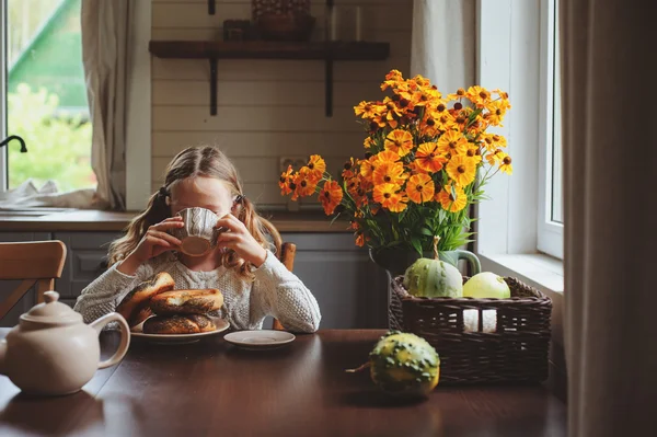 Kind meisje ontbijten thuis in herfst ochtend. Echte leven gezellige moderne interieur in landhuis. Kind bagels eten en het drinken van thee. — Stockfoto