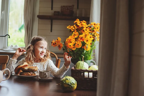 Child girl having breakfast at home in autumn morning. Real life cozy modern interior in country house. Kid eating bagels and drinking tea. — Stock Photo, Image