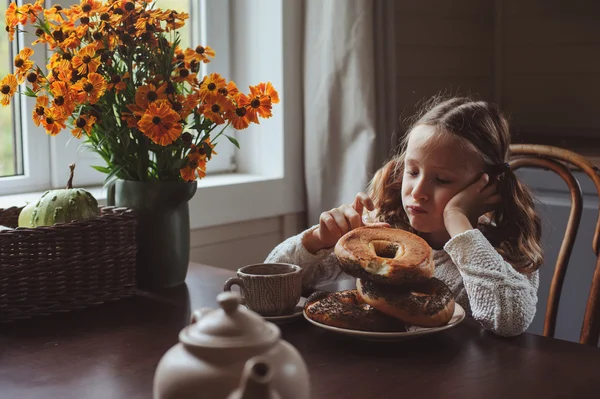 Bambina che fa colazione a casa in autunno mattina. Vita reale accogliente interni moderni in casa di campagna. Bambino mangiare bagel e bere tè . — Foto Stock