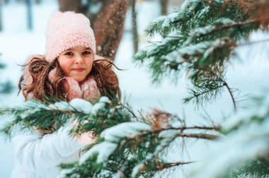Beyaz önlüklü, pembe şapkalı, eldivenli, karlı kış ormanlarında karla kaplı köknar dalıyla oynayan sevimli bir kızın kış portresi. Seyahat, tatil ve aktif kış tatili konsepti