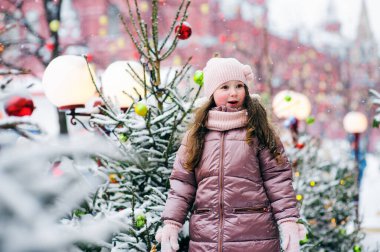 Karlı kış günlerinde dışarıda oynayan mutlu bir kızın Noel portresi. Yeni yıl tatili için süslenmiş köknar ağaçları. Arka planda oyuncaklar ve ışıklar.