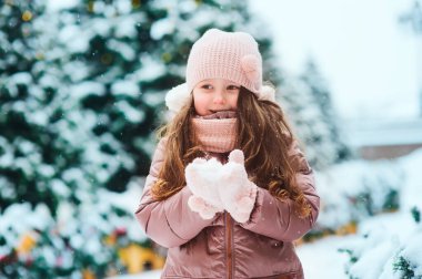 Noel 'de dışarıda oynayan ve kış günü kar atan mutlu bir kızın portresi, köknar ağaçları yeni yıl tatili için süslenmiş oyuncaklar ve arka planda ışıklar.