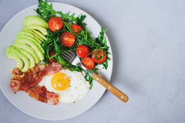Lezzetli kahvaltı - kızarmış yumurta ve domuz pastırması yanında roka salatası ve beyaz tabakta taze avokado.