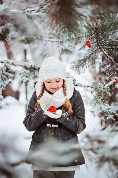 Cute child girl in grey coat and white hat, scarf and gloves plays with toy bullfinch in winter snowy forest — Stock Photo, Image