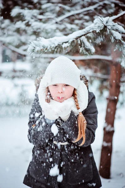 Мила дівчинка в білому капелюсі і шарфі, що дме сніг в зимовому лісі — стокове фото