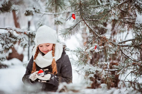 Mignon enfant fille en manteau gris et chapeau blanc, écharpe et gants joue avec jouet bullfinch en hiver forêt enneigée — Photo