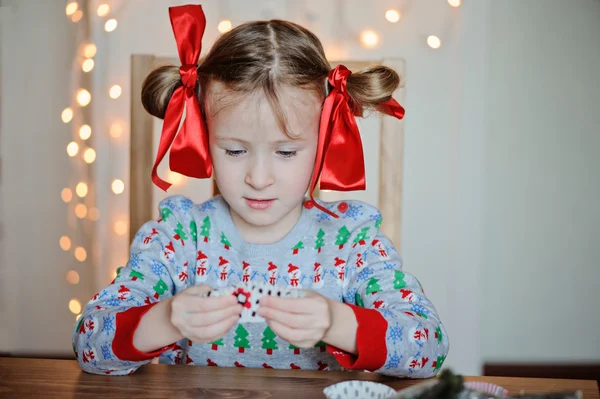 Cute happy child girl in knitted seasonal sweater and with red bows making christmas post cards — Stock Photo, Image