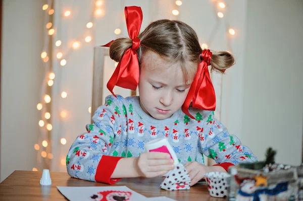 Cute happy child girl in knitted seasonal sweater and with red bows making christmas post cards — Stock Photo, Image