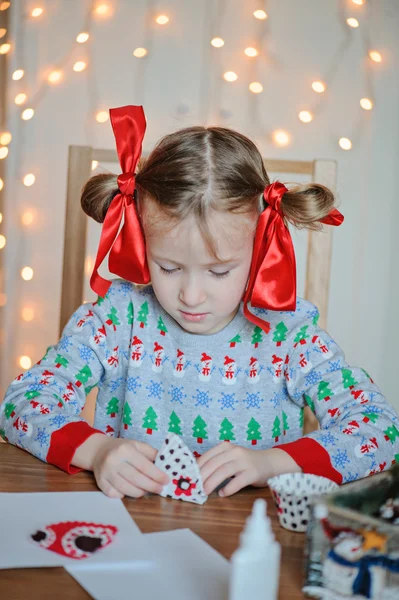 Cute happy child girl in knitted seasonal sweater and with red bows making christmas post cards — Stock Photo, Image