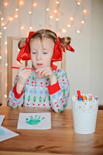 Cute child girl in knitted seasonal sweater with red bows making handprints christmas post cards — Stock Photo, Image