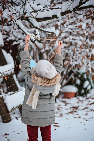 Child girl hangs bird feeder to the tree in winter snowy garden — Stock Photo, Image