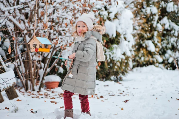 Cute child girl puts seeds to bird feeder in winter snowy garden — Stock Photo, Image