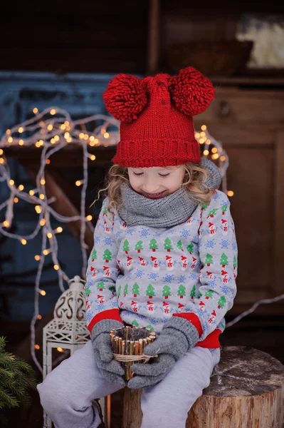 Carino bambino ragazza in maglione di Natale e cappello rosso seduto all'aperto con luci e portacandele in legno — Foto Stock