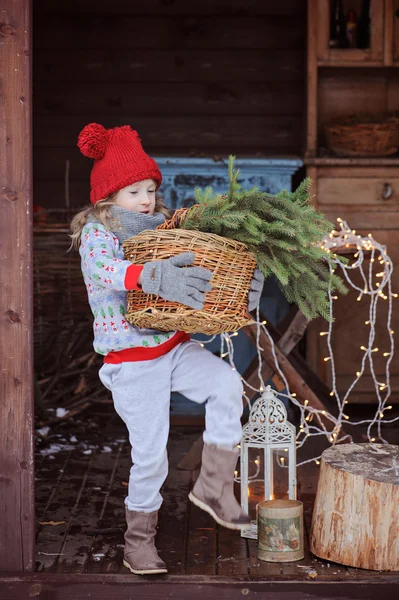 Παιδί κορίτσι σε Χριστούγεννα πουλόβερ και κόκκινο καπέλο κρατώντας το καλάθι των κλάδων της ελάτης — Φωτογραφία Αρχείου
