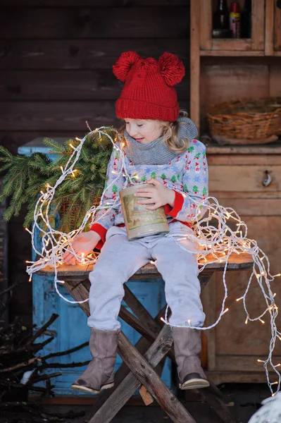 Carino bambino ragazza in maglione di Natale e cappello rosso seduto sul tavolo di legno con scatola di coni con luci sullo sfondo — Foto Stock