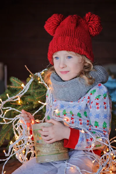 Carino bambino ragazza in maglione di Natale e cappello rosso seduto sul tavolo di legno con scatola di coni con luci sullo sfondo — Foto Stock