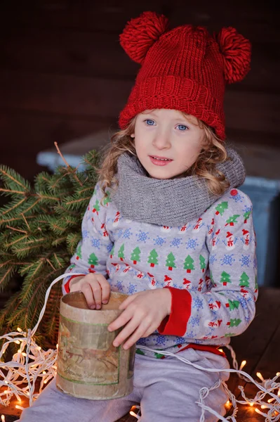 Cute child girl in christmas sweater and red hat sitting on wooden table with box of cones with lights on background — Stock Photo, Image
