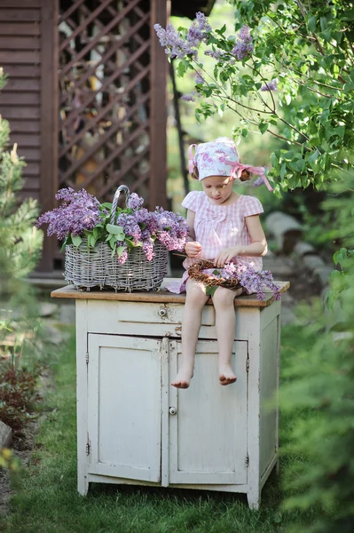 Cute happy child girl in pink plaid dress and flower headband making lilac wreath in spring garden — Stock Photo, Image