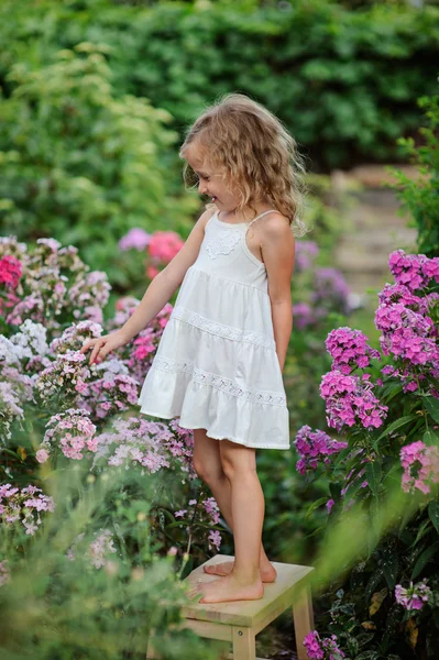 Cute happy blonde child girl in white dress having fun in summer blooming garden — Stock Photo, Image