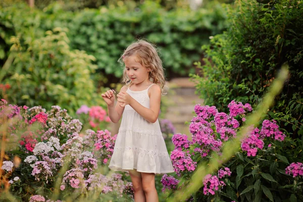 Cute happy blonde child girl in white dress having fun in summer blooming garden — Stock Photo, Image