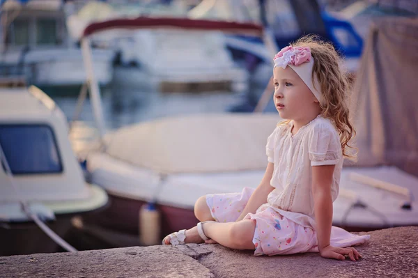 Zonsondergang portret van mooie blonde kind meisje zit aan zee kant in roze en witte outfit met boten op achtergrond — Stockfoto