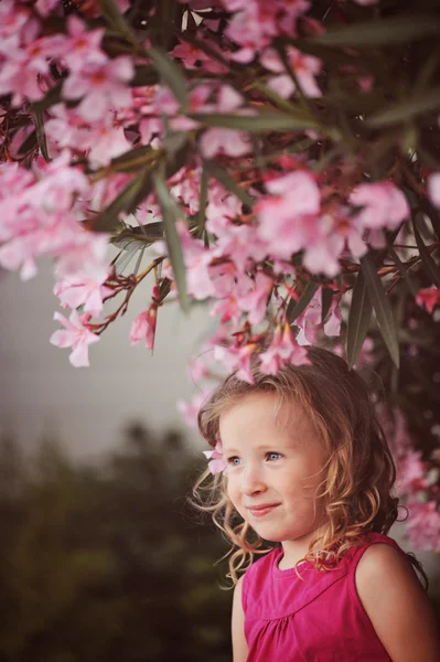 Meisje dat mooie kind zit op de muur onder de roze bloeiende boom in zomerdag — Stockfoto