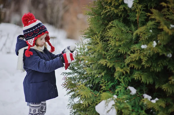 Милая девочка в рождественской шляпе и перчатках, украшающих елку в зимнем саду — стоковое фото
