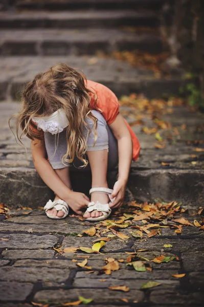 Linda niña en cárdigan naranja jugando y recogiendo hojas mientras está sentada en un viejo camino de piedra con escaleras — Foto de Stock